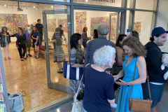 Image of artist opening reception, people gathered inside the gallery looking at the art while people outside talk.