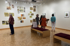 Image from the opening reception, prints are hanging on the walls and a small group of visitors is gathered in the right corner, and a lone visitor is standing on the left of the photograph.