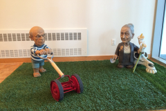 Detail image of Elliott Arkin sculpture that are two of his artist lawn gnomes. On the left is Pablo Picasso mowing a lawn and on the right is Georgia O'Keefe picking flowers. Both sculptures are on a piece of fake grass.