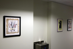 Detail image of artwork hanging in building 12 second floor. This area is known as Farrell Way. Artwork shown by Alan Gordon, an advertisement for a Gorilla Girls exhibition, and an uncut print for a trading card series called "God's Greatest Hits.".