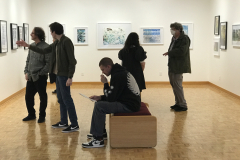 A photograph from Ira Epsteins Exhibition opening. The image shows grous of people around the gallery space, discussing the framed art work hanging on the walls.