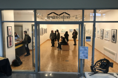 A photograph from Ira Epsteins Exhibition opening. The image shows grous of people around the gallery space, discussing the framed art work hanging on the walls.