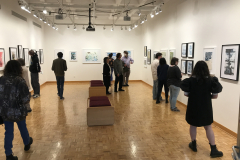 A photograph from Ira Epsteins Exhibition opening. The image shows grous of people around the gallery space, discussing the framed art work hanging on the walls.
