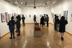 A photograph from Ira Epsteins Exhibition opening. The image shows grous of people around the gallery space, discussing the framed art work hanging on the walls.