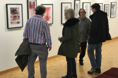 A photograph from Ira Epsteins Exhibition opening. The image shows grous of people around the gallery space, discussing the framed art work hanging on the walls.