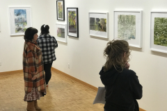 A photograph from Ira Epsteins Exhibition opening. The image shows grous of people around the gallery space, discussing the framed art work hanging on the walls.