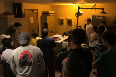 Image from Jenn Libby's Wet Collodion Demonstration. This image shows Jenn in the background of a darkroom pouring chemicals on a plate, and many students around her watching.
