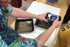 Image from Jenn Libby's Wet Collodion Demonstration. This image shows a close up of the image in the photographic tray, and a close up of a pair of hands taking a photograph of the image is in the foreground.