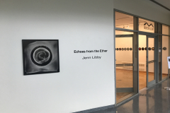 Installation image of Jenn Libby exhibition Echoes from the Ether. This image is from the outside of the gallery, with a large black and white abstract photograph on the left of the image, text announcing the show in the center, and on the right we can see into the gallery, showing the right wall and a row of seven inch albums in a row.