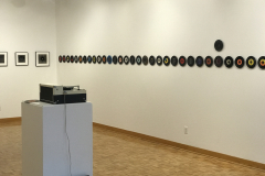 Installation image of Jenn Libby exhibition Echoes from the Ether.  This image shows a detail of the right wall, which contains a row of seven inch albums lined up very closely. A singular album stands out above the rest.. A small area of the back wall can be seen, showing smaller black and white abstract images. In the foreground is an old record player.