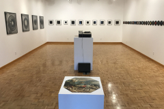Installation image of Jenn Libby exhibition Echoes from the Ether. This image is looking into the back of the gallery, with large black and white abstract photographs on the left wall, smaller black and white abstract photographs on the back wall, and small seven inch records with very small black and white images in the center lining the right wall. In the foreground is an old record player and a puzzle.