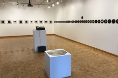 Installation image of Jenn Libby exhibition Echoes from the Ether.  This image shows a detail of the right wall, which contains a row of seven inch albums lined up very closely. A singular album stands out above the rest.. A small area of the back wall can be seen, showing smaller black and white abstract images. In the foreground is an old record player and a octagonal puzzle showing a detail of a Bruegel painting.