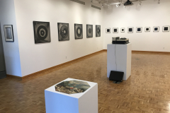 Installation image of Jenn Libby exhibition Echoes from the Ether.  This image shows the left wall containing large black and white abstract images. A small area of the back wall can be seen, showing smaller black and white abstract images. In the foreground is an old record player and a octagonal puzzle showing a detail of a Bruegel painting.