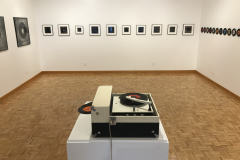 Installation image of Jenn Libby exhibition Echoes from the Ether. This image is looking into the back of the gallery, with large black and white abstract photographs on the left wall, smaller black and white abstract photographs on the back wall, and small seven inch records with very small black and white images in the center lining the right wall. In the foreground is an old record player.