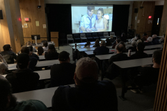 A group of people sit in a darkened room, all watching the screen at the front of the room showing a film.