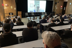 A group of people sit in a darkened room, all watching the screen at the front of the room showing a film.