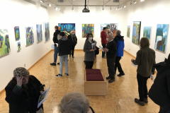 Photograph from the opening reception for Judy Gohringer. This image is shot from above and shows a group of visitors looking at the art work on the wall, some people are talking with others.