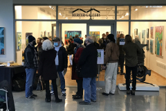 Photograph from the opening reception for Judy Gohringer. This image is taken from outside the gallery, and shows a group of visitors outside the gallery talking, and inside the gallery there are many people looking at the artwork.