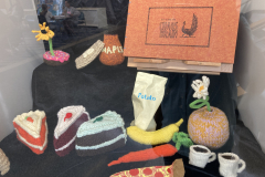 An image of the display case, showing various handmade objects, including a piece of pizza, some pieces of cake, and a book. The objects are made of yarn or paper mache.