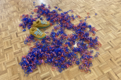 A close up of the floor sculpture, showing a handmade pair of shoes and a large amount of plastic strips.
