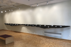 Installation shot of right wall, including long angled and curved shelf with soil on it, and in the soil are many ceramic oval pieces containing war images of planes, parachutes, and bombs.