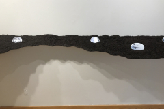 Detail of the Installation of right wall, including long angled and curved shelf with soil on it, and in the soil are many ceramic oval pieces containing war images of planes, parachutes, and bombs.