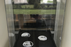 Installation shot of a pedistal, with soil and three ceramic ovals containing war imagery. Behind the pedestal is a window with a printed cling attached, and it is of a landscape and the horizon line matches up with the horizon behind it.