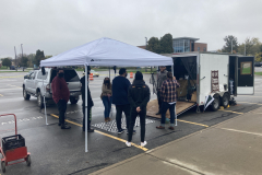 A photograph taken out doors, to the right is a trailer with photographic darkroom equipment in it, and to the left is a pop up tent with a group of people standing under it.