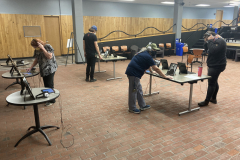 An installation shot of the exhibition, many tables are spread out, and on the tables are framed photographs and iPads with headphones. At different tables we see four people looking at the work and listening to the audio interviews.