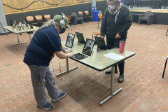 An installation shot of the exhibition, many tables are spread out, and on the tables are framed photographs and iPads with headphones. There are two people at one of the tables looking at the work and listening to the audio interviews.