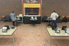 An installation shot of the exhibition, many tables are spread out, and on the tables are framed photographs and iPads with headphones. At different tables we see two people looking at the work and listening to the audio interviews.