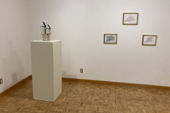 Installation photography of Nate Hodge Exhibition. In the image we see  a small sculpture of a house on a pedestal on the left and three small framed drawings on the right.