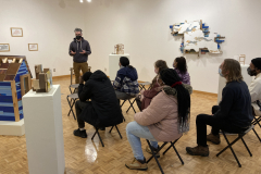 A photograph of Nate Hodge lecturing about his work to a group of people.  This photograph was taken in the gallery so his work is featured around the people, the artist is standing, and the guests are sitting on chairs.