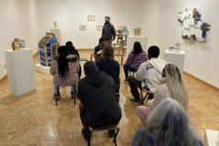 A photograph of Nate Hodge lecturing about his work to a group of people.  This photograph was taken in the gallery so his work is featured around the people, the artist is standing, and the guests are sitting on chairs.