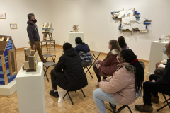 A photograph of Nate Hodge lecturing about his work to a group of people.  This photograph was taken in the gallery so his work is featured around the people, the artist is standing, and the guests are sitting on chairs.
