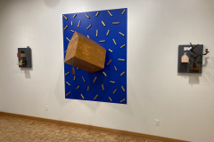 Detail image of the Robert Booth gallery installation, showing a detail of the rear wall. The left and right sculptures are made of small houses and branches mounted on slate. The slate has hash marks or couds drawn on in chalk. The center piece is a larger house protruding from the wall that has been painted in a bright blue color. In the field of blue there are many small branches that have been painted gold.