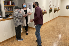 An image of the opening reception for Robert Booth. In this image we see three people standing and talking, and in the background we can see a part of the  gallery installation, including four wall sculptures.