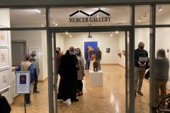 An image of the opening reception for Robert Booth. In this image we see the gallery from the atrium outside. Inside the gallery are a number of people talking amongst all the artwork.