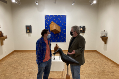 An image of the opening reception for Robert Booth. In this image we see a closeup of the artist talking with someone, standing next to a floor sculpture.