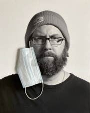 A close photograph of a bearded mad wearing a knit hat and black t-shirt. Attached to the image is a paper mask that appears to hang from the right ear of the person.