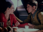 A still image from a film.  A distressed looking woman in a red sweater sitting at a breakfast table, touching the wrist of a stoic man in a yellow sweater. The man is looking into her eyes and touching her shoulder. The breakfast table has empty plates, cups, and a plate of fruit. Bright light is filtering into the room from behind them.